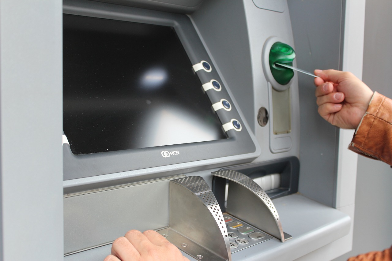 Close-up of a person using an ATM. The ATM is light gray metal. A person's hand, wearing a light brown or tan-colored jacket, is inserting a credit card into the card reader slot, which has a green button. The ATM screen displays a black background. The keypad for entering a PIN is visible, as well as a slot for receiving cash dispensed. The ATM's exterior is clean and modern.