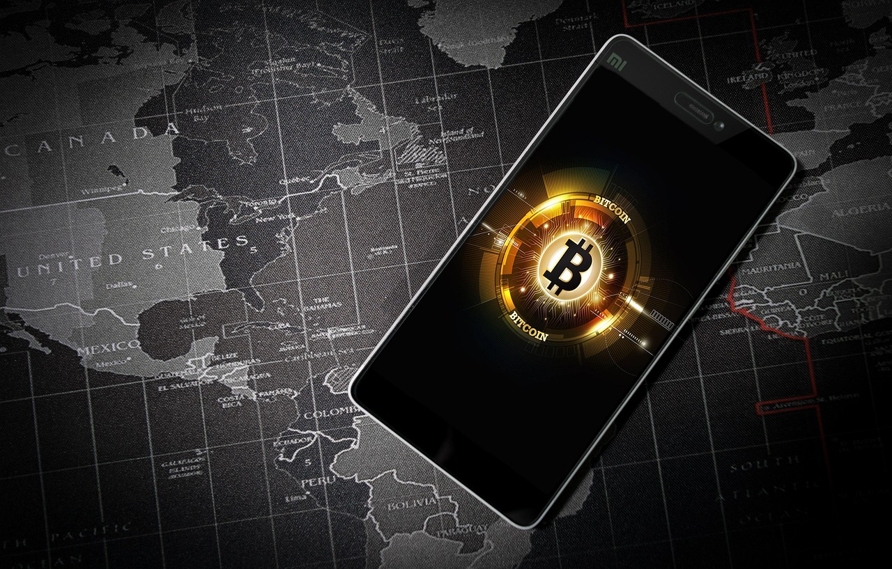A smartphone, positioned diagonally across a dark-colored world map, displays a graphic of the Bitcoin symbol.
            The map, in shades of gray and black, is detailed with country outlines.