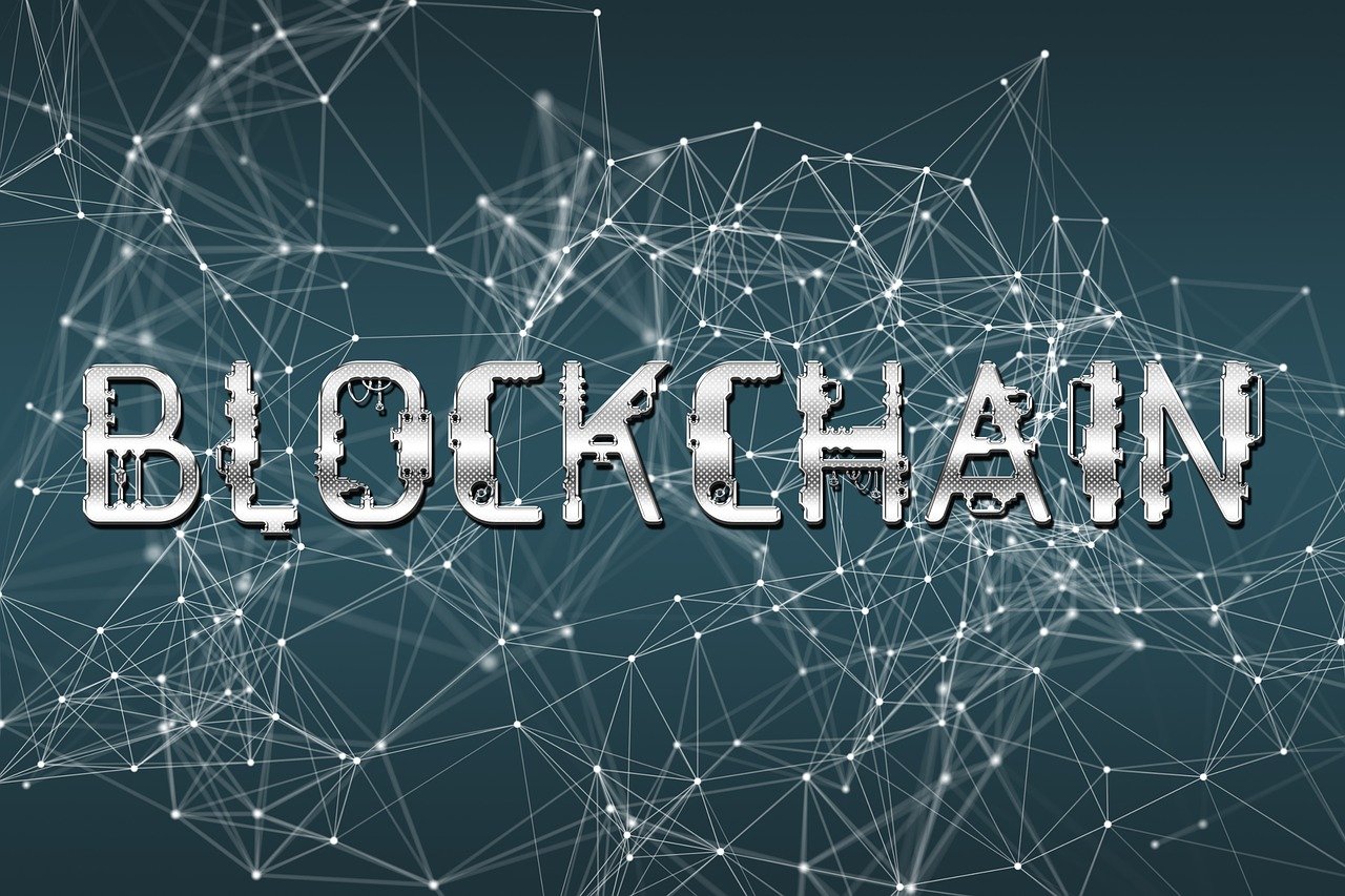 The image features the word "BLOCKCHAIN" rendered in a stylized, metallic silver font. The letters have a slightly distressed or fragmented look, giving the impression of a circuit board or mechanical design. The background is a dark teal or navy blue color. A complex network of interconnected lines and dots, resembling a network or digital connection, covers the background. The lines and dots are a light gray or white, creating a sense of depth and complexity behind the text. The overall effect is a modern, technological portrayal of the concept of blockchain.